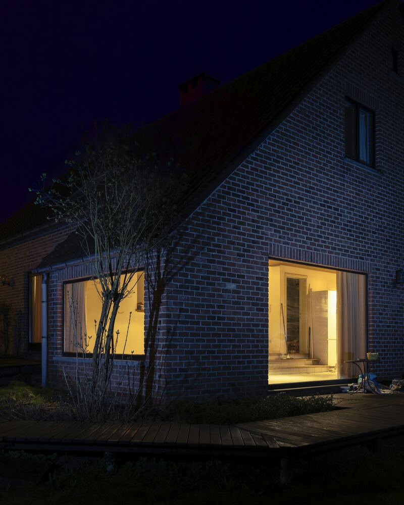 Maison en brique la nuit, deux grandes fenêtres éclairées de l’intérieur.