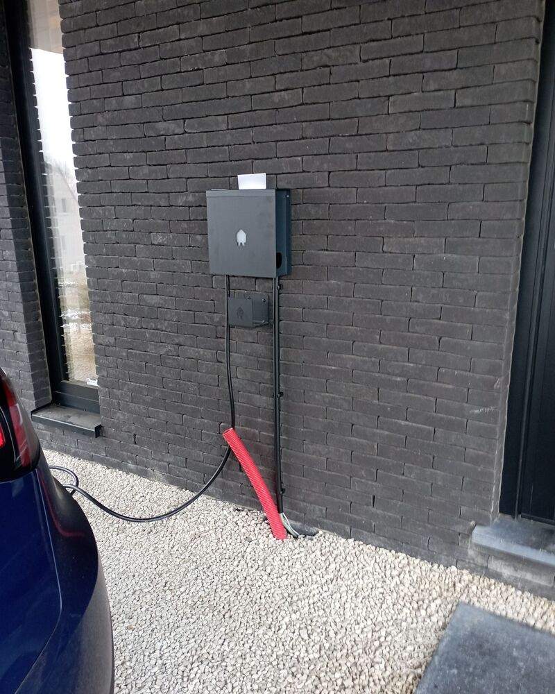 Borne de recharge murale fixée sur une façade en brique sombre, câble raccordé à une voiture.